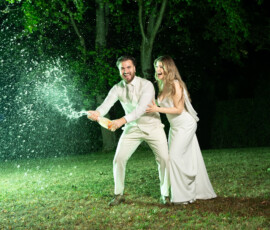 Braut und Bräutigam öffnen bei Nacht eine Champagnerflasche auf einer Wiese. Die Champagnerfontäne wird mit professioneller Blitzlicht-Fotografie eingefangen. Exklusive Hochzeitsfotografie und Hochzeitsvideografie in Stuttgart von Majestic Wedding.